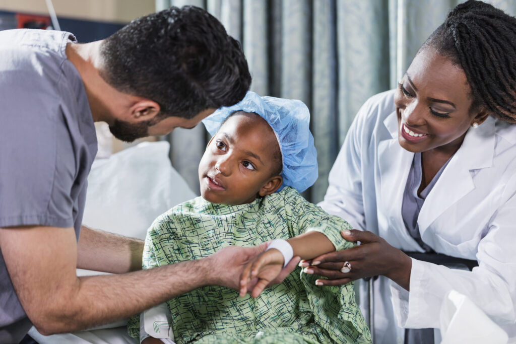 A 6 year old African-American girl sitting on a hospital gurney, with a doctor and nurse tending to her.  She is wearing a hospital gown and surgical cap. A nurse in scrubs, a young Middle Eastern man in his 20s, is standing on one side of her, and a female doctor, a mature African-American woman in her 40s, is standing on the other side.